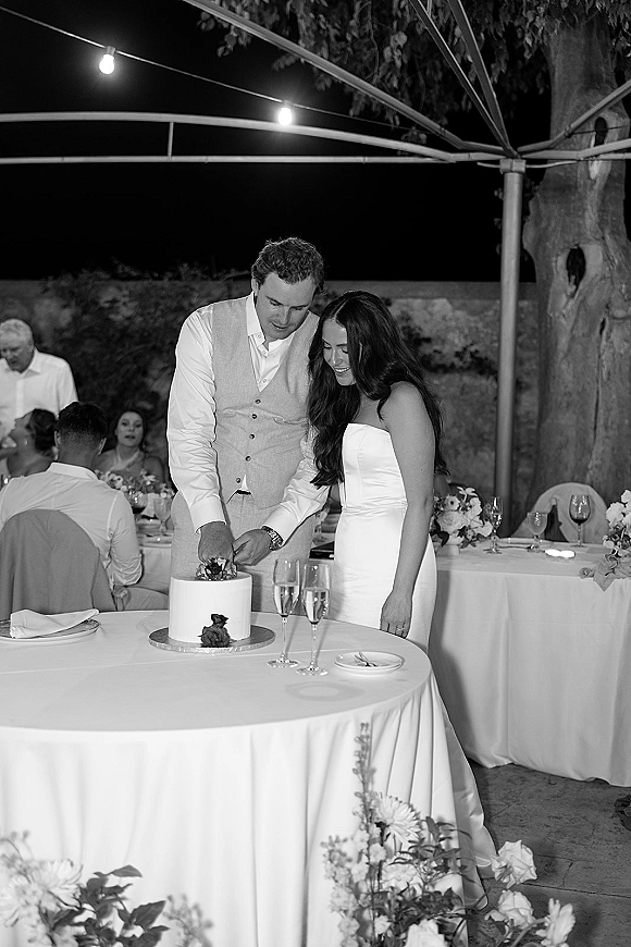 Wedding cake cutting as bride in a strapless dress and groom in vest slice a white cake under string lights at an outdoor night reception