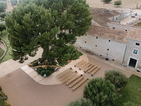 Outdoor ceremony setup with wood chairs lining a paved courtyard aisle, floral markers and boulders beside a stone building under a pine tree