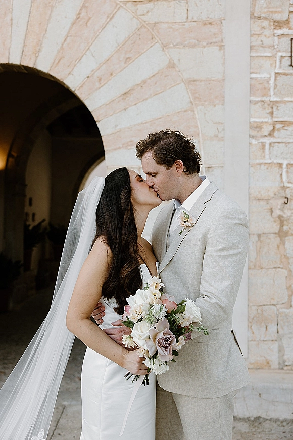 Wedding kiss portrait of bride and groom kissing under a stone archway, bride in strapless dress holding bouquet, groom in light gray suit