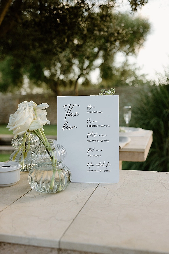 Wedding bar menu and signature drinks sign displayed on a bar counter with white roses in clear glass vases on a stone patio