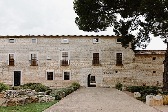 Wedding venue exterior with a stone facade and arched doorway, featuring wood doors and balcony railings by a driveway and lawn under cloudy sky