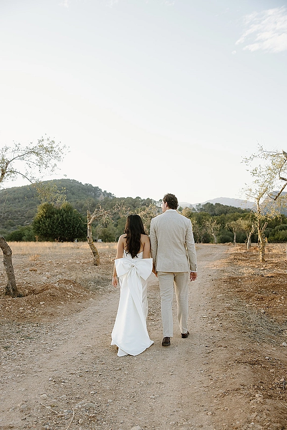 Couple portrait of bride and groom walking away hand in hand, her strapless gown with oversized bow, on a dirt road with mountains behind