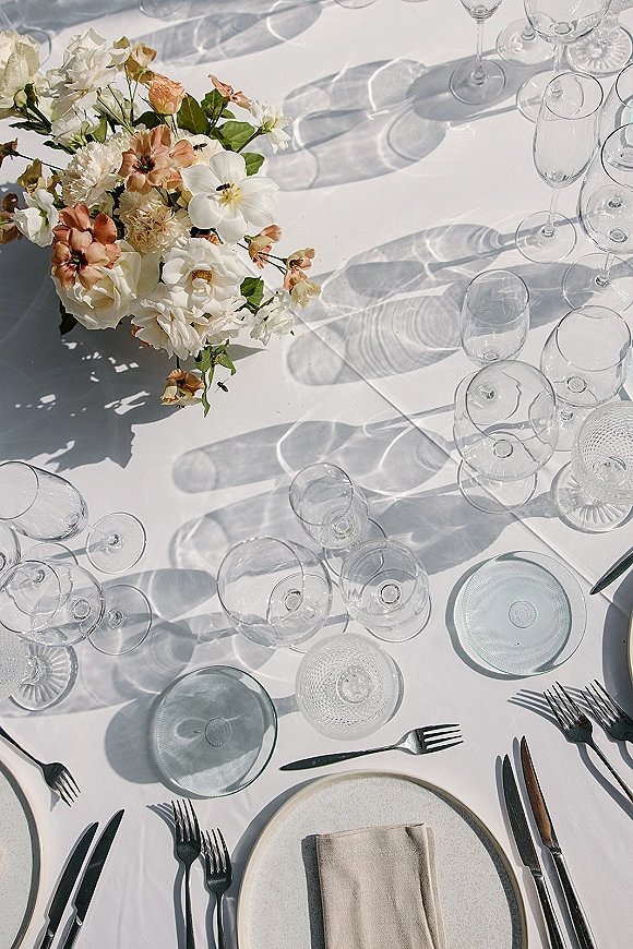Reception tablescape with a wedding table setting on a white tablecloth, floral centerpiece, glass plates, and wine glasses in sunlight shadows