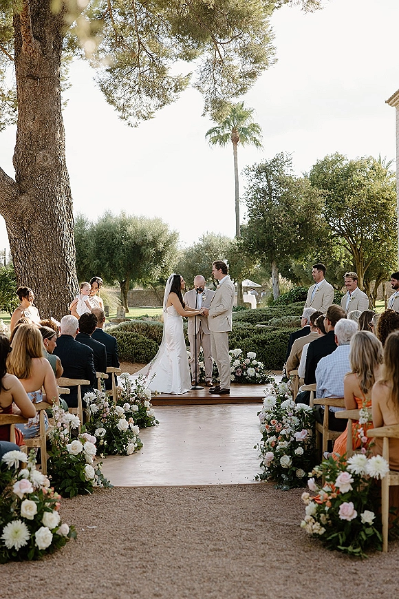 Wedding vows at an outdoor wedding ceremony as bride in long veil and groom in beige suit speak at a microphone under a floral arch in a garden aisle