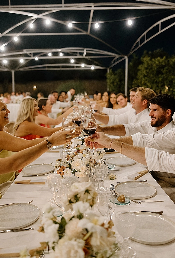 Wedding reception toast as guests clink wine glasses and champagne flutes over a long white tablescape under string lights in a tent at night