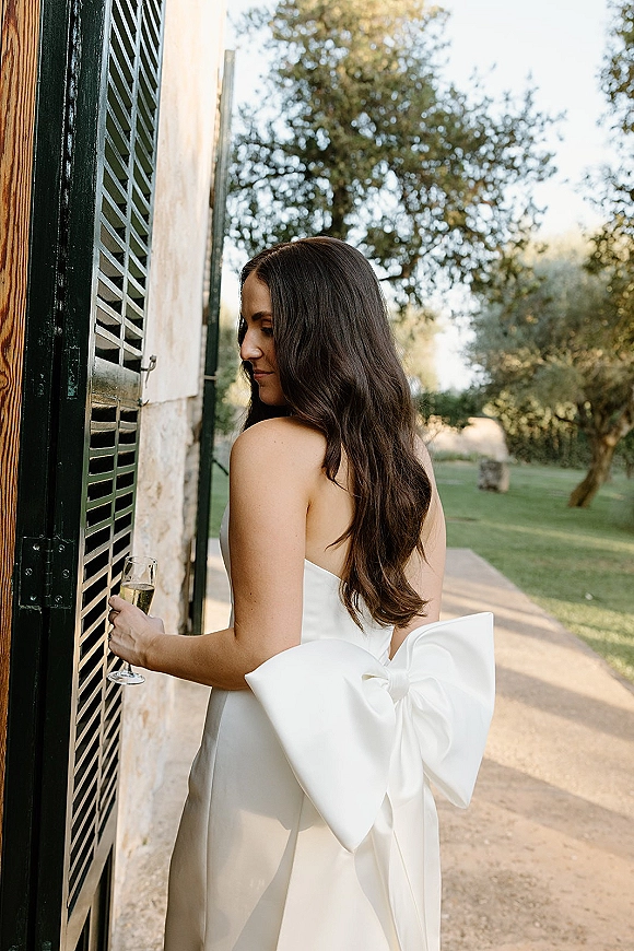 Bridal portrait of a bride in a strapless wedding dress with an oversized bow, holding a champagne flute by stone wall and shutters in daylight