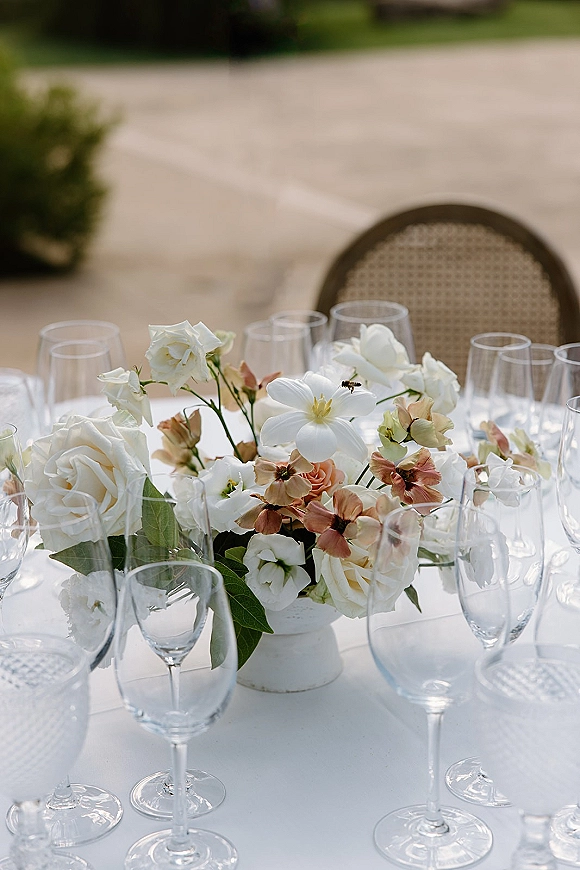 Wedding centerpiece with a white floral centerpiece in a white vase, surrounded by wine glasses on a white tablecloth on an outdoor patio lawn