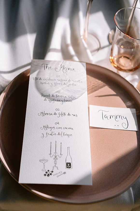 Wedding place setting with a wedding menu card in calligraphy atop a charger plate, plus place card, napkin, and glass on white linen with sun shadows