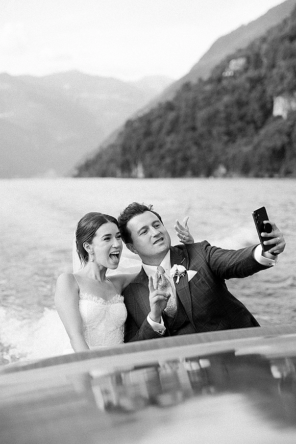 Wedding couple selfie as the bride in veil and drop earrings laughs beside groom in suit on a boat railing with mountain lake behind