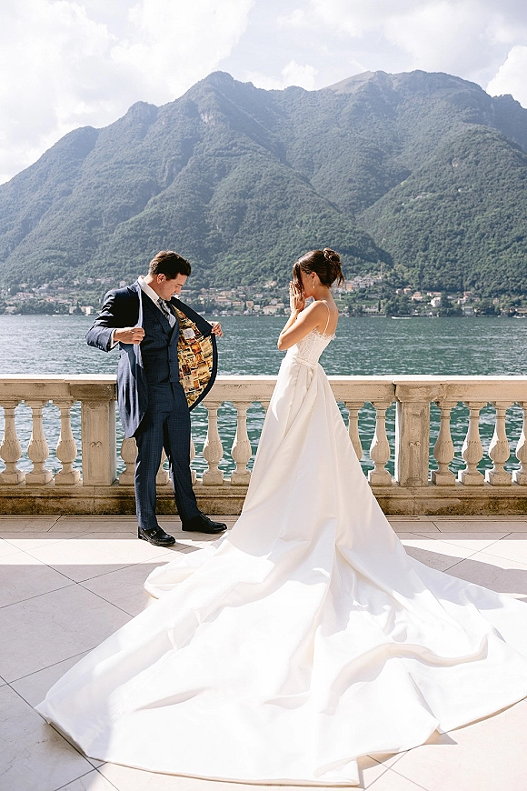 First look moment on a lakefront terrace as the groom reveals his jacket lining and the bride covers her face, mountains beyond under clouds