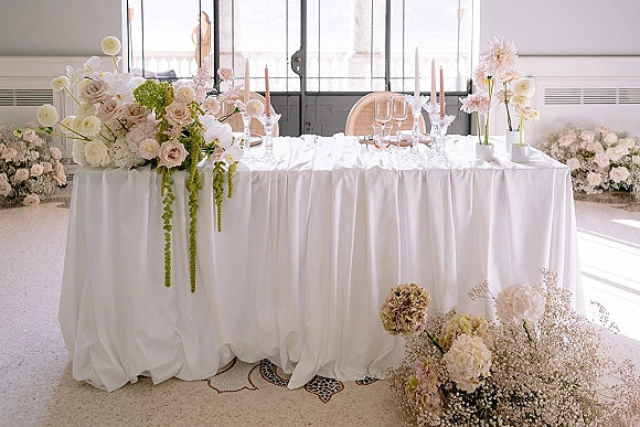 Reception sweetheart table with floral arrangements and taper candles on a white tablecloth, set by large windows in soft natural light