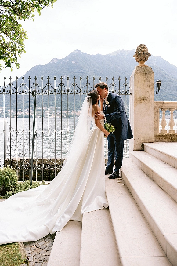 Wedding kiss portrait of bride and groom kissing, her cathedral veil trailing on stone steps by an iron gate with lake and mountains beyond