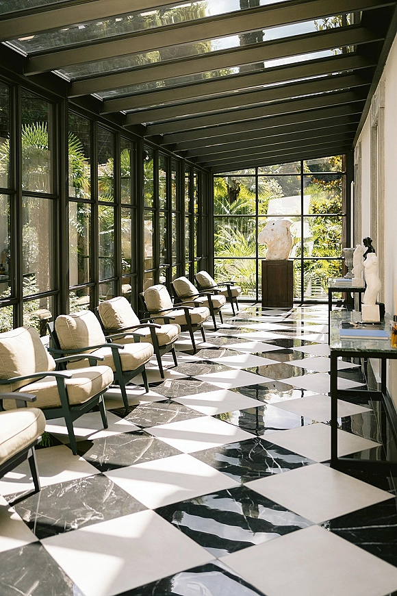 Venue lounge area with wedding venue lounge seating, upholstered chairs beside side tables and books in a sunlit glass-roof sunroom with palms