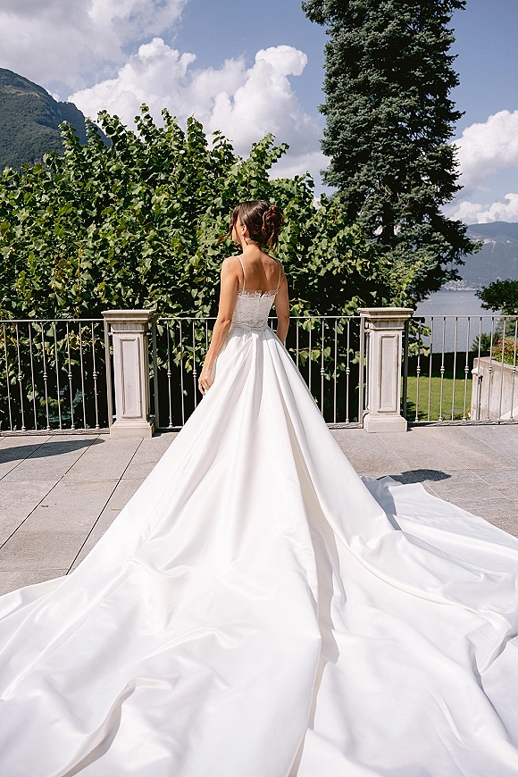 Bridal portrait of a bride in a back view wedding dress with a long train on a stone terrace overlooking mountains and a lake under blue sky