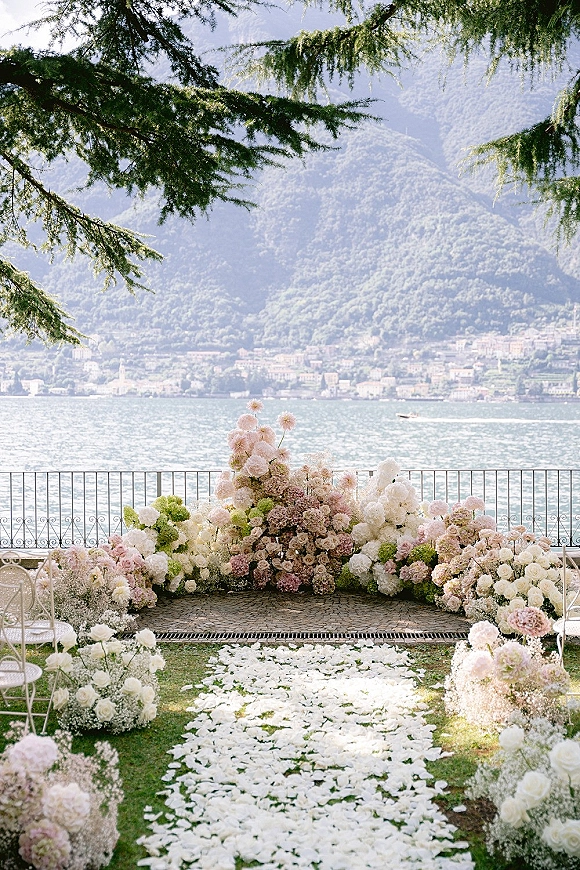 Ceremony setup for an outdoor wedding ceremony with floral arch, blush and white blooms, and white rose petal aisle by a lake with mountains