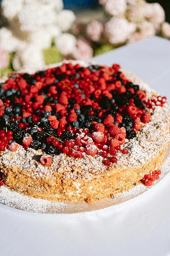 Wedding cake topped with mixed berries and powdered sugar on a cake stand, set on a white tablecloth with blurred florals behind