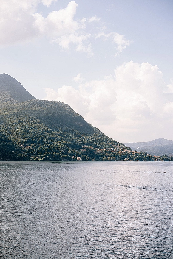 Lake landscape with calm blue-gray water facing mountains and a forested hillside, with a lakeshore village beneath a cloudy sky