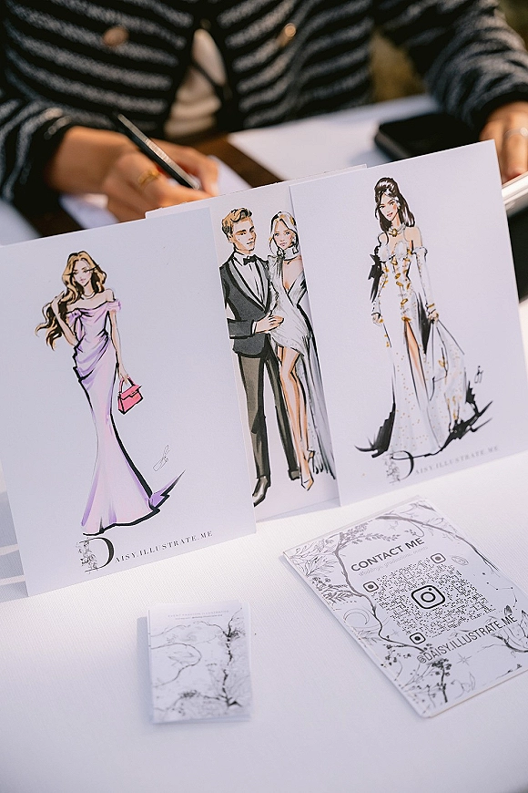 Live wedding illustrator setting up a vendor table with fashion illustration prints, sketch cards, pen, and QR code sign on a tablecloth