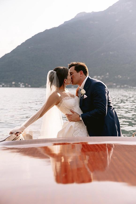 Wedding kiss as newlyweds on boat embrace, bride in lace dress and veil holding groom’s face on a lake with mountain backdrop