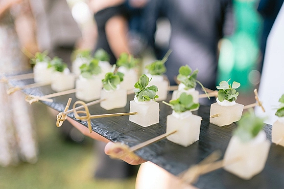 Wedding cocktail appetizers on a slate serving tray with skewered cheese cubes and microgreens, blurred guests in an outdoor setting