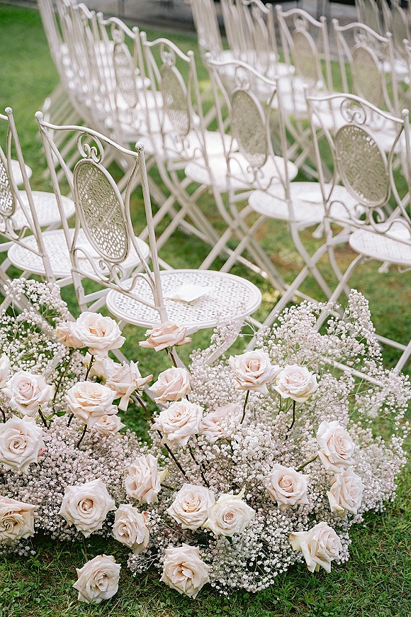 Ceremony seating with outdoor ceremony chairs in rows of white metal chairs, blush roses and baby’s breath lining a grass garden aisle