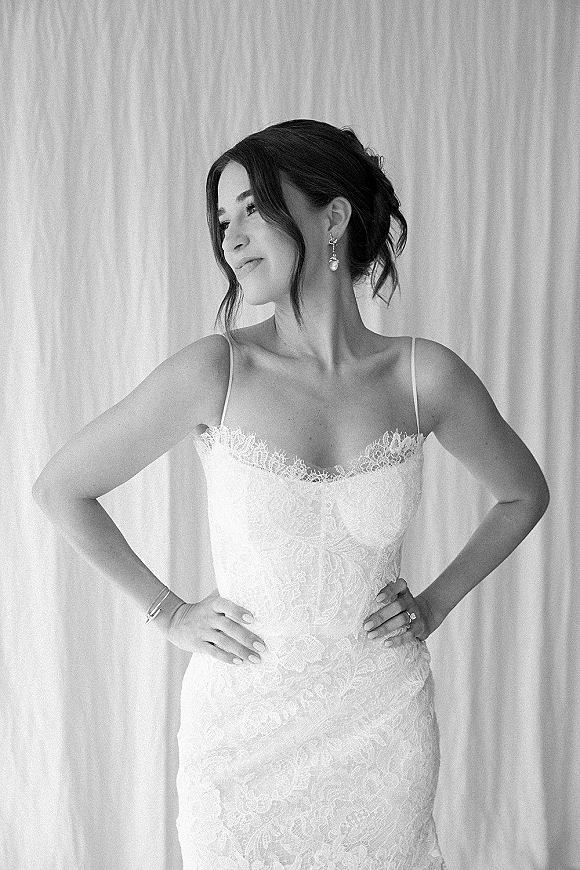 Bridal portrait in black and white of a bride with hands on hips, lace spaghetti-strap gown, drop earrings, and updo before a curtain backdrop