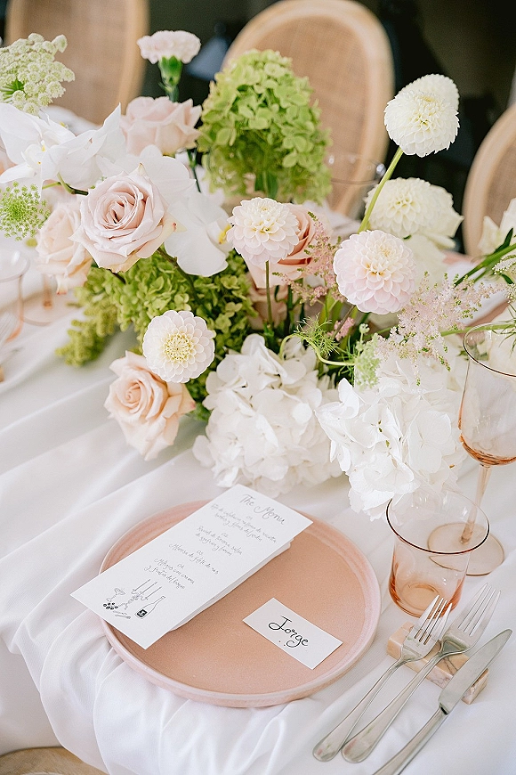 Wedding tablescape with a hydrangea centerpiece, blush charger plates, rose-gold rim glassware, and menu cards on a white tablecloth reception table