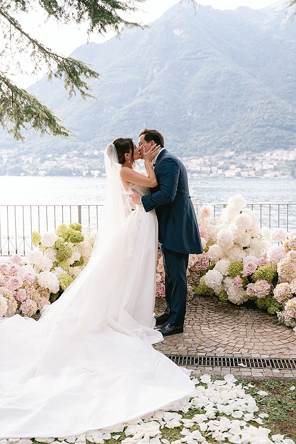 Wedding kiss portrait of bride and groom kissing, her veil and long train flowing on a lakeside terrace with mountain backdrop