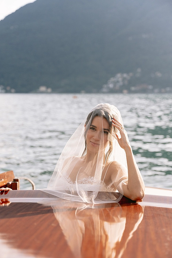 Bridal portrait of a bride in veil on a wooden boat, wearing a strapless lace dress and ring, with lake and mountains behind