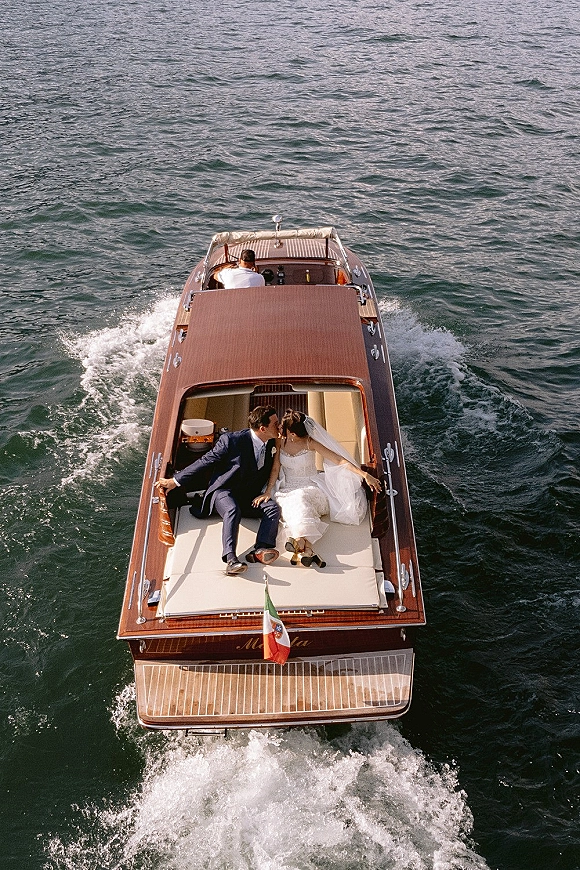 Wedding couple portrait of bride and groom on boat kissing, her long veil trailing as a motorboat cuts through open water wake