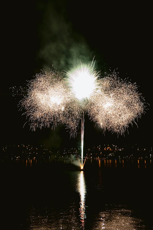 Wedding fireworks burst over a lake at night, golden sparks and smoke mirrored in the water with shoreline lights glowing in the distance