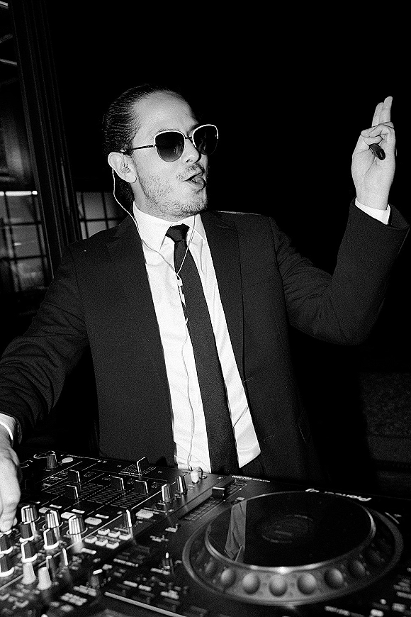 Wedding DJ in sunglasses and suit working a DJ controller with headphones wire at a dark indoor reception with window frames