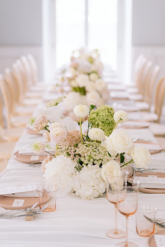 Reception tablescape with wedding table centerpiece of white roses, hydrangea and dahlias, blush napkins, menu cards, amber stemware by bright windows