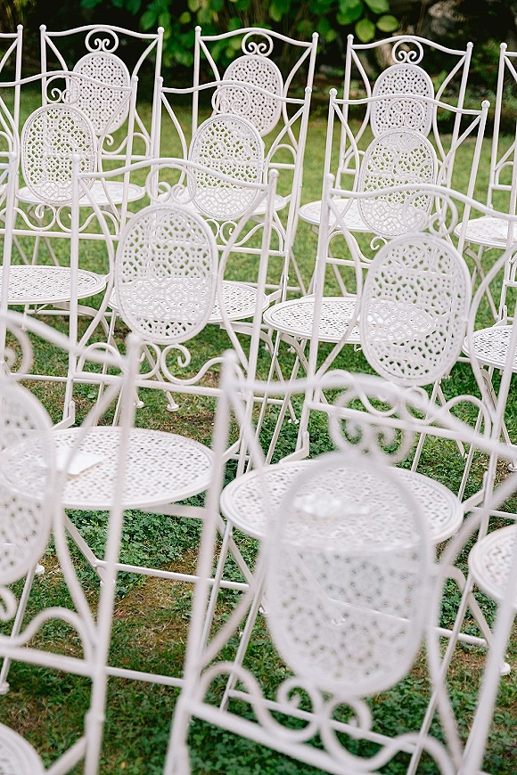 Ceremony seating with white wrought iron chairs arranged on a grass lawn, surrounded by lush garden foliage for an outdoor wedding setup