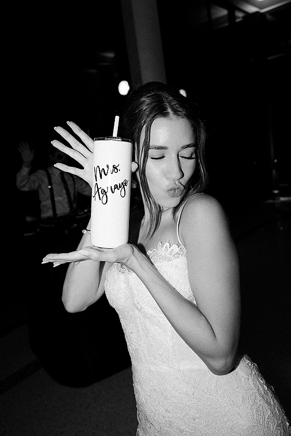 Bride portrait in a black and white wedding photo, holding a personalized tumbler with straw and ring, posing on a dark reception dance floor