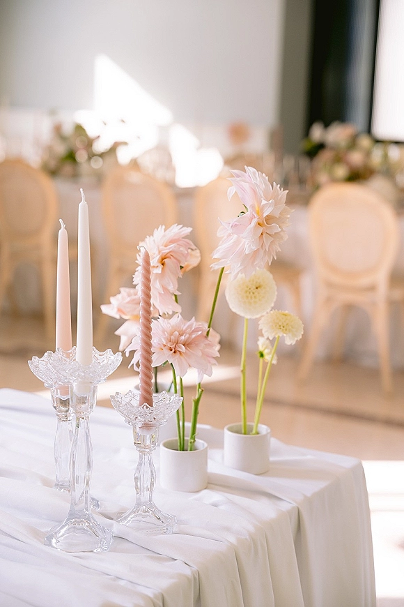Wedding centerpiece with pastel dahlias in bud vases and glass candlesticks with taper candles on a white tablecloth, window-lit reception tables