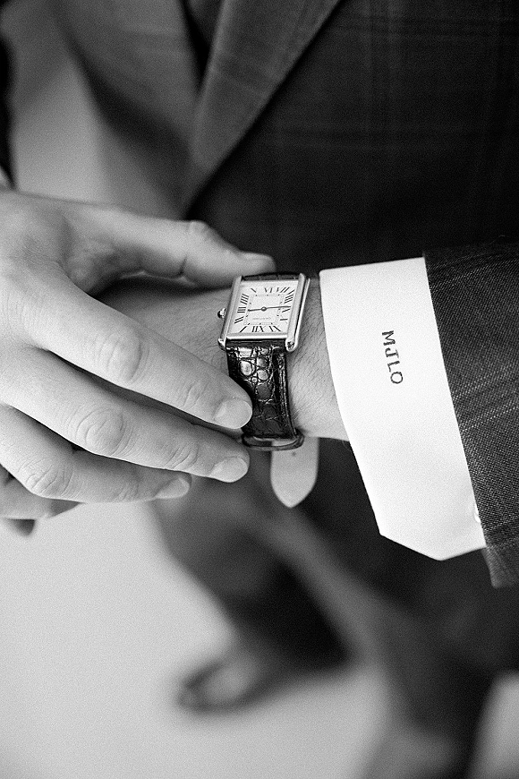 Groom accessories with a wedding watch detail as a hand adjusts a rectangular dress watch against a suit cuff in a blurred indoor setting