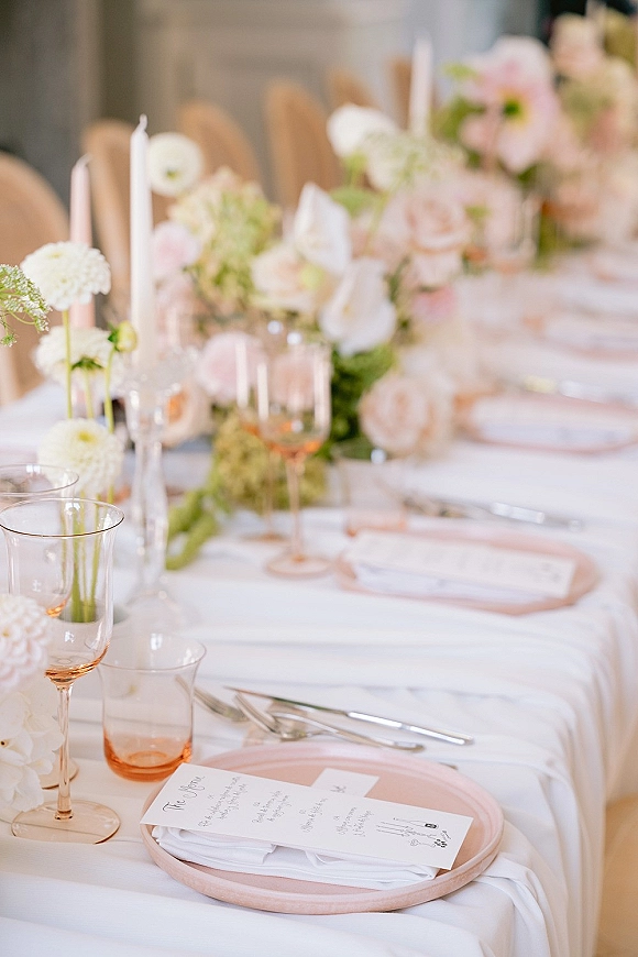 Wedding tablescape with wedding place setting featuring pink and white floral centerpieces, white taper candles, menus on plates, and glassware on linens
