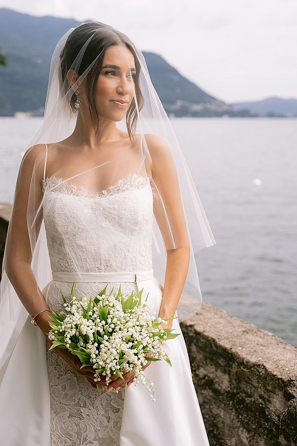 Bridal portrait of a bride holding bouquet with a flowing veil, lace gown, and lily of the valley by a mountain lake stone wall