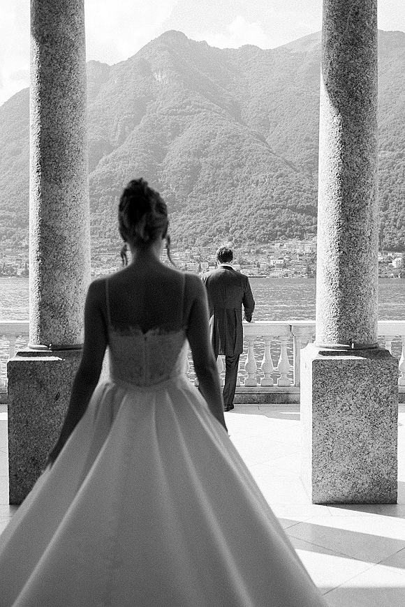 Wedding first look as bride approaches groom from behind, lace bodice and tulle skirt flowing on a terrace with lake and mountains beyond