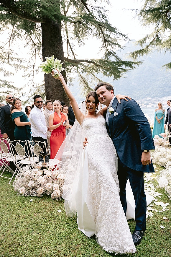 Recessional moment as bride raises bouquet beside groom in blue suit, walking past cheering guests on rose petal aisle by lake and mountains