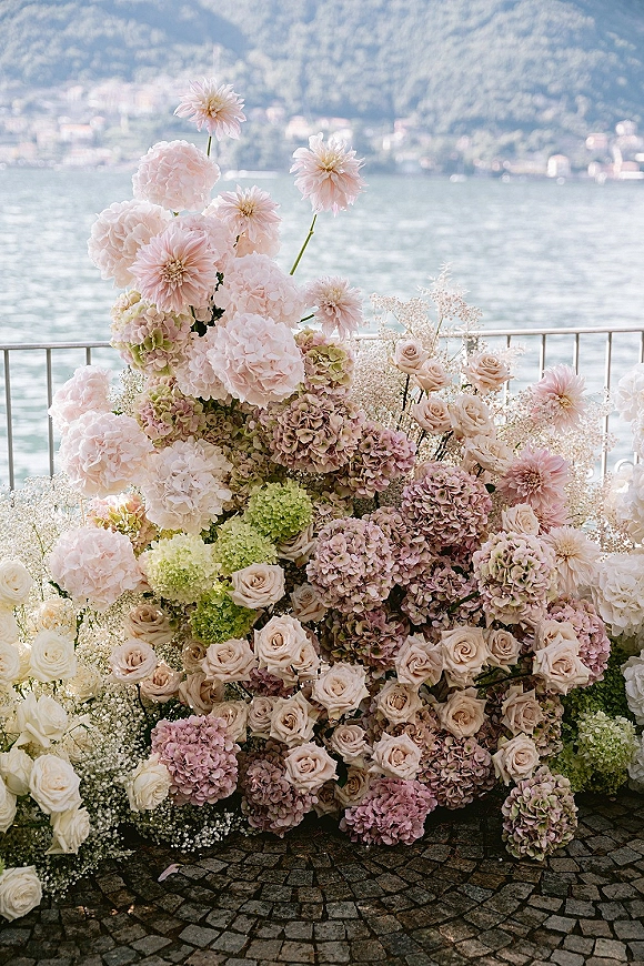 Wedding floral arrangement with hydrangeas and roses in soft pastels beside a metal railing, with lakeside water and mountains behind