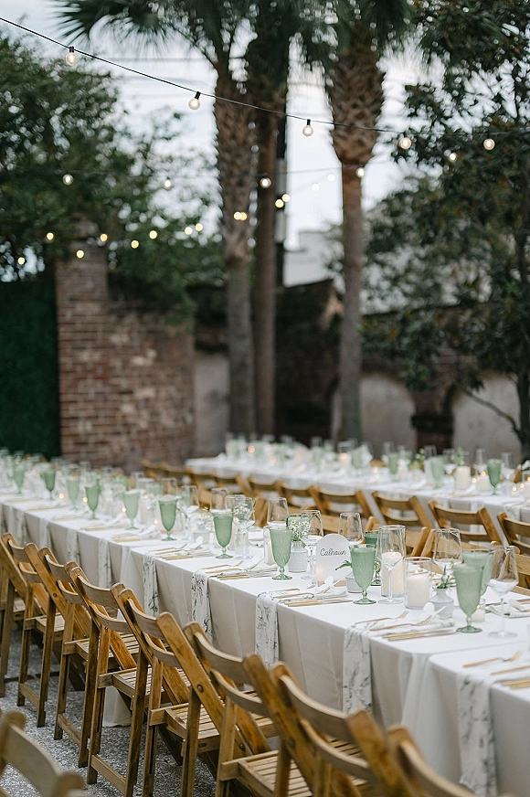 Reception tablescape with long banquet table in a courtyard, marble runners, green goblets, taper candles, and string lights at dusk