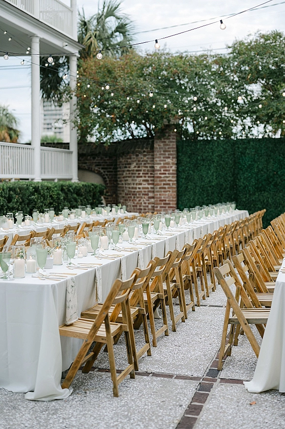 Reception tablescape with long banquet table setup, white linens, wooden folding chairs, votive candles and string lights in a courtyard patio