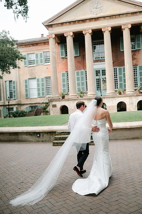 Couple portrait of bride and groom walking away, cathedral veil trailing over her train in a brick courtyard by columned building