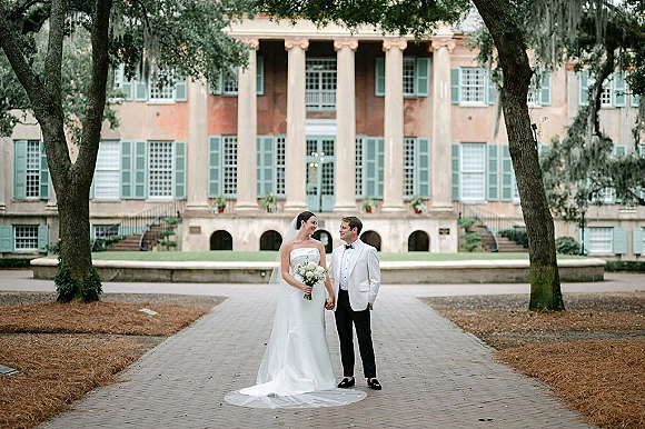 Couple portrait of bride and groom holding hands, bride with veil and white bouquet, by columned estate on brick path with mossy trees