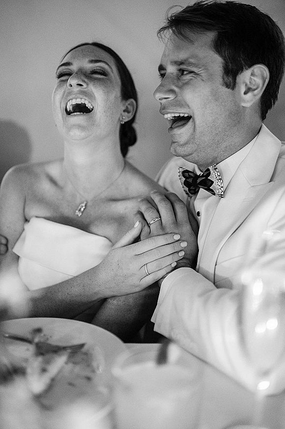 Reception candid moment of bride and groom laughing at their table, holding hands, with wedding rings visible and plates in front