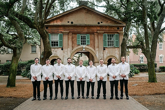 Groomsmen portrait of a lineup in white dinner jackets and bow ties, standing on a brick walkway before a historic arched doorway