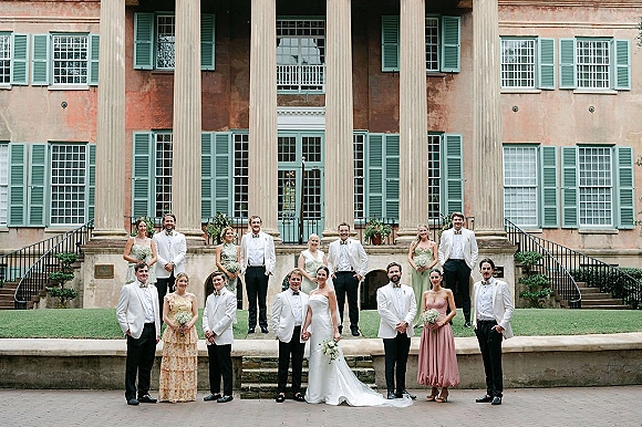 Wedding party portrait with bride and groom lined up in formal attire, bouquets in hand, on the front steps of a historic mansion with columns