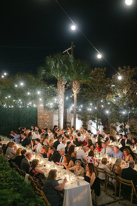 Outdoor wedding reception with string lights over long banquet tables, candlelit white linens, greenery, and wooden chairs in a palm-lined courtyard at night